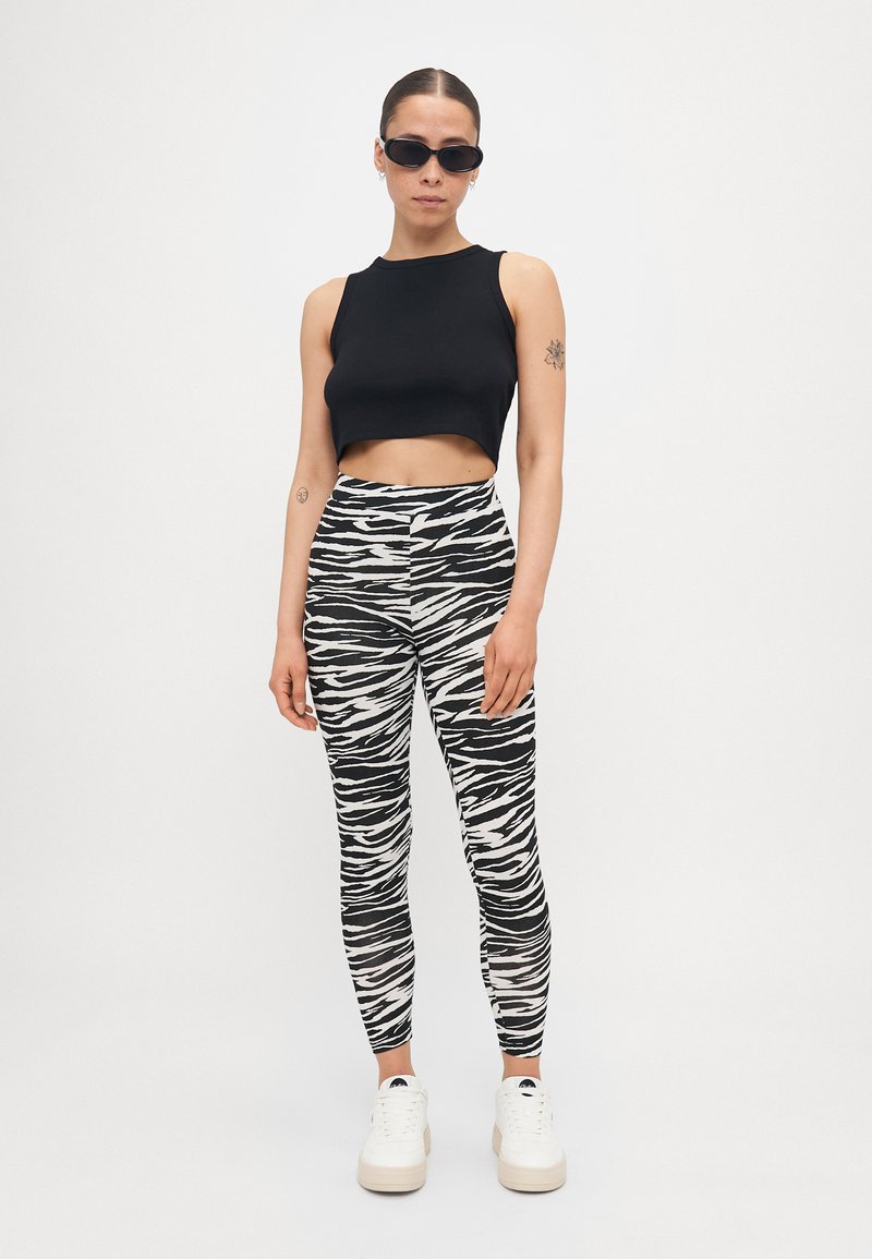 Fekete cropped tank top, fekete-fehér zebra mintás leggings és vastag talpú fehér sportcipő. A modellen fekete napszemüveg van.