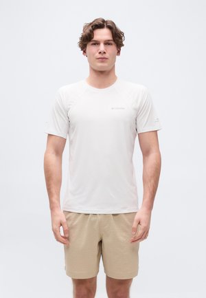 Jeune homme aux cheveux courts et ondulés portant une chemise blanche Columbia à manches courtes et un short beige texturé, debout devant un fond uni.