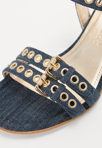 Denim hæl-sandaler med guldfarvede nitter og spænder, en blød inderside og en mørkeblå struktureret yderside.