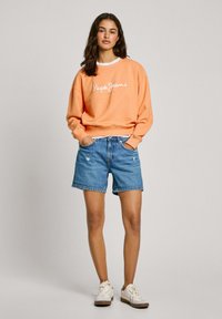 Sweatshirt orange avec logo blanc, associé à un short en denim bleu. Le mannequin porte des baskets blanches. Il dispose de manches longues et d'une coupe décontractée.