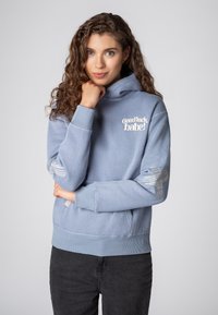 Lichtblauwe hoodie met een voorkeurszak, ribgebreide boorden en zoom. Voorzien van witte tekst "Good luck, babe!" en zilveren gestreepte accenten op de mouwen.