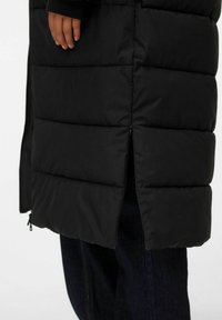 Melna pūkaina jaka ar vertikāliem quiltētiem sekcijām, sānu rāvējslēdzēja detaļām un augstu apkakli. Audumam ir gluda faktūra, nodrošinot siltumu un komfortu.