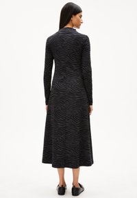 Femme avec de longs cheveux noirs portant une robe midi zèbre noire et grise et des chaussures noires à enfiler, tournée de dos sur un fond blanc.