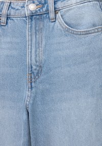 Jean en denim bleu clair en gros plan montrant la taille, le bouton, les passants de ceinture, la poche avant et les coutures détaillées sur le tissu.