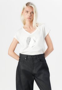 T-shirt blanc à manches courtes avec un design de cœur texturé argenté sur le devant, associé à un pantalon en denim noir taille haute.
