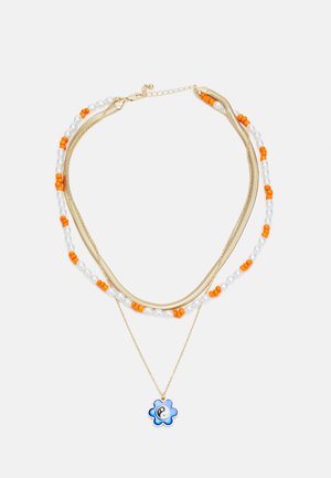 Colar em camadas em tom dourado, apresentando contas laranja, pérolas brancas e um pendente em forma de flor azul com um símbolo yin-yang.