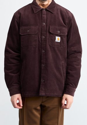 Homme portant une chemise en velours côtelé marron foncé à boutons avec deux poches poitrine, dont une montrant un patch logo Carhartt, et un pantalon marron.