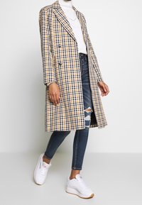 Abrigo de doble botonadura en un patrón de cuadros amarillo, negro y blanco. Presenta un cuello, mangas largas y una textura suave. Llevado sobre vaqueros y zapatillas.