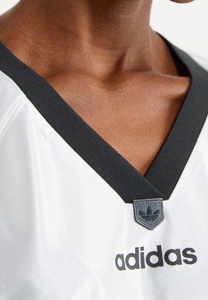 Osoba o ciemnej karnacji ubrana w białą koszulkę Adidas z czarnym dekoltem w serek i naszywką z logo na środku szyi.