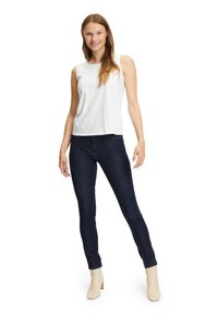 Giovane donna in piedi, con una maglietta bianca senza maniche, jeans blu scuri e stivali alla caviglia beige, che sorride con i lunghi capelli castano chiaro sciolti.