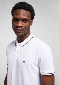 Uomo con capelli corti e ricci che indossa una polo bianca Lee con rifiniture nere, che guarda leggermente verso la fotocamera su uno sfondo semplice.