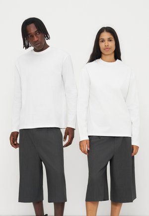 Deux modèles portant des hauts blancs à manches longues et des shorts gris foncé amples jusqu'aux genoux, debout devant un fond clair uni.
