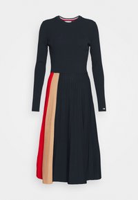 Gebreide jurk met lange mouwen, donkerblauw bovenstuk, geplooide rok in zwarte, rode en beige secties, ronde halslijn, textiel met structuur, gouden accenten.