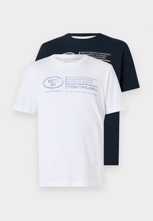 Weißes T-Shirt mit kurzen Ärmeln und blauem Aufdruck, kombiniert mit grauen Jeans. Das Model steht lässig mit den Händen in den Taschen und trägt braune Schuhe.