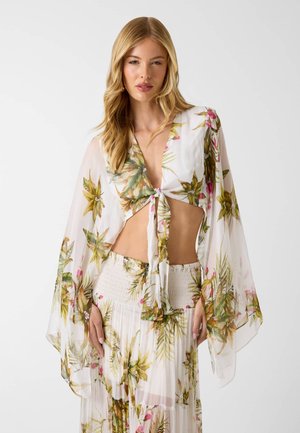 Femme blonde portant un haut floral blanc transparent noué à l'avant et une jupe florale taille haute assortie sur fond blanc.