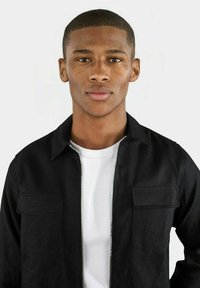 Svart zip-up jacka med två framfickor över en vit T-shirt. Jackan har krage och en slät, texturerad tyg.