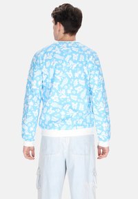 Hanorac albastru deschis cu un model floral și de frunze alb. Manșete și tiv ribbate albe. Modelul poartă pantaloni cargo de culoare deschisă.