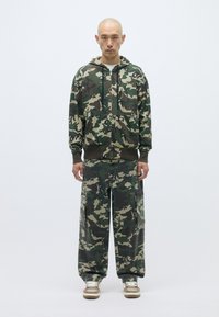 Kameras kapuci un saskaņotas cargo bikses. Kokvilnas maisījums, zaļš un brūns raksts, priekšējās kabatas kapucē un biksēs, brīvā piegriezuma, ar regulējamu kapuci.