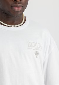 Camiseta de algodón blanca con texto bordado en gris claro "BERLÍN ALEMANIA" y un gráfico de un globo aerostático. Cuello redondo, corte relajado.