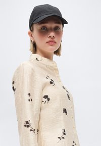 Νέα γυναίκα που φοράει μαύρο καπέλο και μπέζ πουκάμισο με μαύρα λουλουδένια σχέδια, κοιτάζει με αυτοπεποίθηση την κάμερα μπροστά σε απλό φόντο.