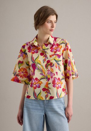 Femme portant une chemise à manches courtes avec des motifs floraux lumineux et un jean bleu clair, regardant vers sa gauche sur un fond uni.