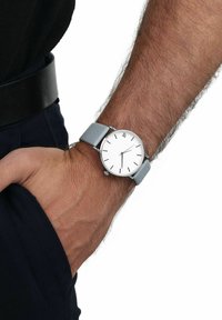 Runder Silberne Uhr mit weißem Ziffernblatt, schwarzen Stundenmarkierungen und grauem Lederarmband, das am Handgelenk getragen wird. Mit einer Krone an der Seite versehen.