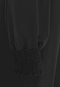 Svart plaggdetalj som visar en rynkad, elastisk mudd med en jämn yta och en subtil glans på tyget.