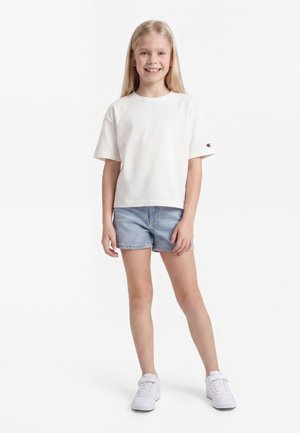 Ung pige med langt blondt hår iført en hvid t-shirt, denimshorts og hvide sneakers, smilende og stående mod en ensfarvet baggrund.