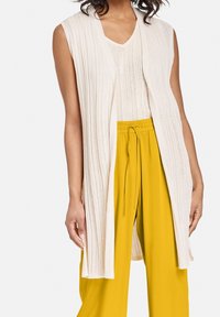 Gilet lungo e aperto davanti in tessuto crema testurizzato con design a coste, indossato su un top coordinato e pantaloni gialli a vita alta.