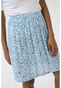 Frau in einem hellblauen Blumenkleid mit elastischem Bund und einem weißen eng anliegenden Kurzarmoberteil, die mit entspannten Händen steht.