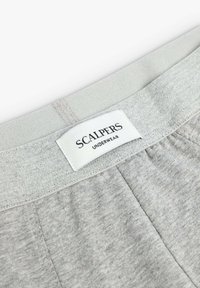 La ropa interior gris presenta una tela suave y texturizada con una cinturilla elástica. La etiqueta dice "SCALPERS UNDERWEAR" en negro sobre fondo blanco.