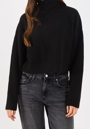 Femme portant un pull à col roulé noir avec poignets et ourlet côtelés, associé à un jean noir délavé taille haute.