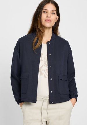 Woman with long brown hair wearing a navy buttoned jacket over a light shirt with a beige abstract design, beige pants, hands in pockets.
