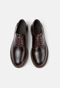 Une paire de chaussures de ville à lacets en cuir poli marron foncé vue de dessus sur un fond clair.