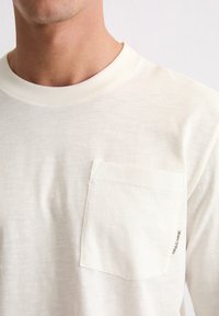 Nahaufnahme eines weißen Rundhalsshirts mit einer Brusttasche und einem kleinen Stoffetikett am Rand der Tasche.