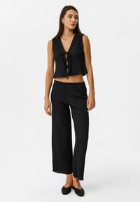 Zwarte, gestructureerde crop top met een diepe V-hals en veterdetail aan de voorkant, gedragen met wijde, zwarte broek met elastische taille en platte zwarte schoenen.