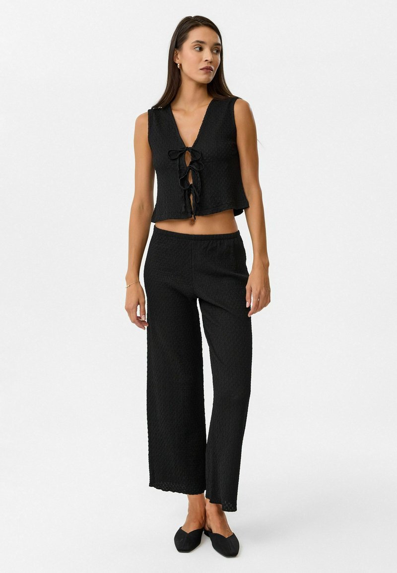 Zwarte, gestructureerde crop top met een diepe V-hals en veterdetail aan de voorkant, gedragen met wijde, zwarte broek met elastische taille en platte zwarte schoenen.
