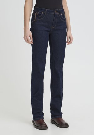 Mujer con jeans azul oscuro de pierna recta, blusa negra y botas de tobillo marrón oscuro, de pie contra un fondo liso y claro.