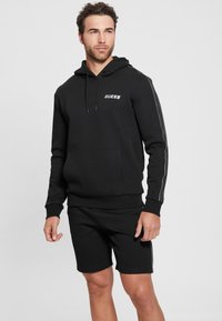 Black hoodie with white logo on chest, featuring a drawstring hood and side stripes. Paired with matching black shorts. Smooth texture.