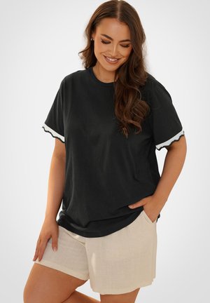 Kvinde med langt brunt hår iført en sort kortærmet top med hvide kanter og beige shorts, smilende og kigger nedad med den ene hånd i lommen.