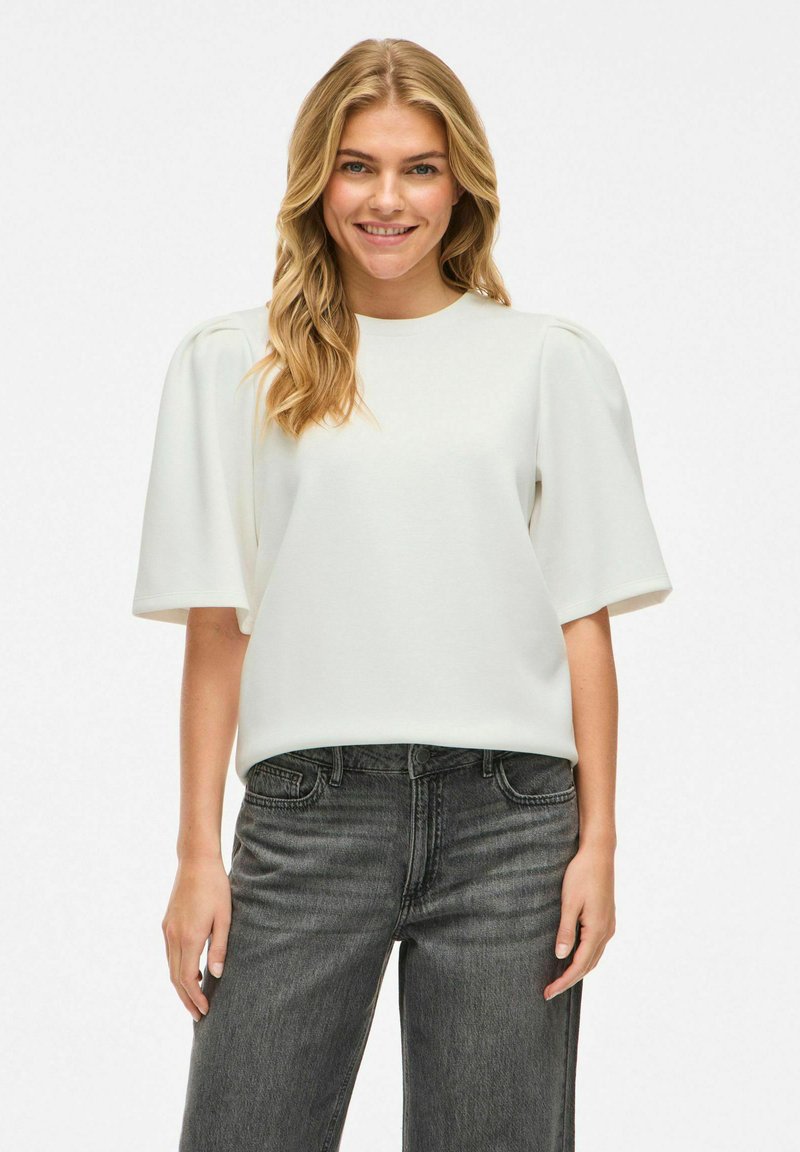 Pull blanc à manches courtes bouffantes. Tissu doux et lisse. Associé à un jean taille haute gris foncé. Design simple et moderne.