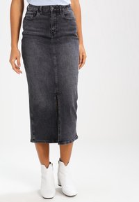 Falda midi de mezclilla negra con una abertura frontal. Cuenta con cinco bolsillos, cintura ajustada y una silueta recta clásica. Se lleva con botines blancos.