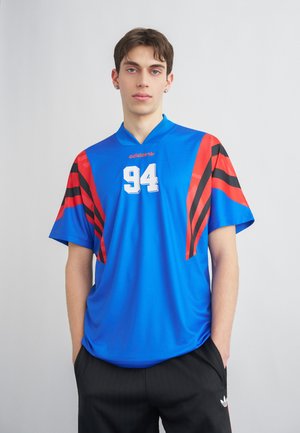 Νέος άνδρας που φοράει μπλε αθλητική μπλούζα Adidas με κόκκινες και μαύρες ρίγες και αριθμό 94, στέκεται με τα χέρια στις τσέπες του μαύρου παντελονιού.