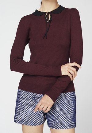 Pull à manches longues couleur marron avec un col noir et un détail de cravate, associé à un short en maille bleu marine avec un motif géométrique.