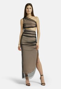 Ein trägerloses, durchsichtiges Kleid in taupe mit schwarzen Rüschen-Details, hohem Schlitz und seitlichem Ausschnitt. Kombiniert mit schwarzen, schnüreartigen Absätzen.