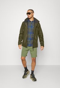 Veste imperméable vert olive sur une chemise à carreaux grise, assortie à un short vert clair et des baskets marron avec des accents bleus. Grandes lunettes de soleil.