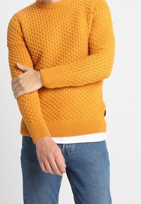 Homme portant un pull orange texturé et un jean bleu, croisant un bras pour tenir le coude opposé, sur un fond uni.