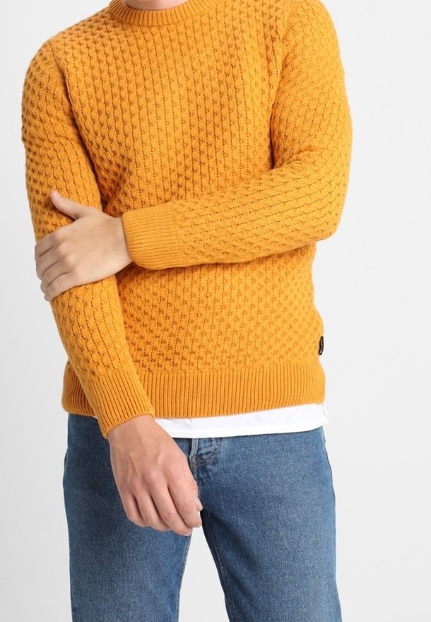 Homme portant un pull orange texturé et un jean bleu, croisant un bras pour tenir le coude opposé, sur un fond uni.