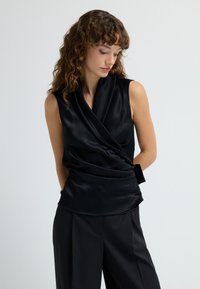 Top negro sin mangas en material satinado con un diseño drapeado. Presenta un escote en V profundo y una textura suave, combinado con pantalones negros a medida.