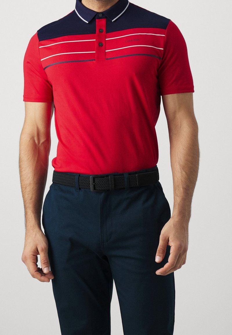 Červené polo tričko s námořnicky modrým límcem, s vodorovnými pruhy v bílé a námořnicky modré barvě. Nosené s tmavě námořnickými kalhotami a černým tkaným pásem.